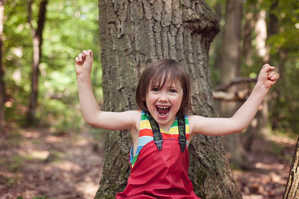 Child wearing red waterproof rain pants in wooded outdoor setting, laughing with arms raised